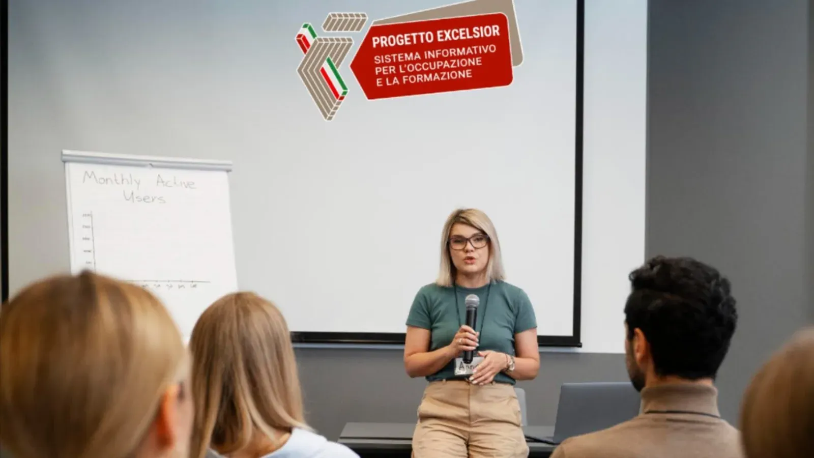 Relatrice con microfono davanti a una platea durante un incontro, con il logo Progetto Excelsior